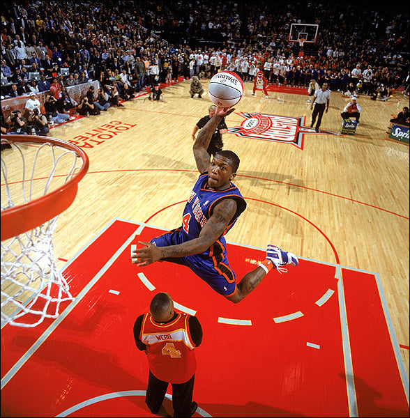 A basketball player in a blue and orange uniform dunks over another player during a crowded NBA event, with spectators and a red court visible in the background.