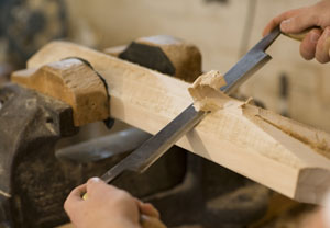 Close-up of hands using a metal file to shape a piece of wood secured in a vise, with wood shavings being removed from the surface.