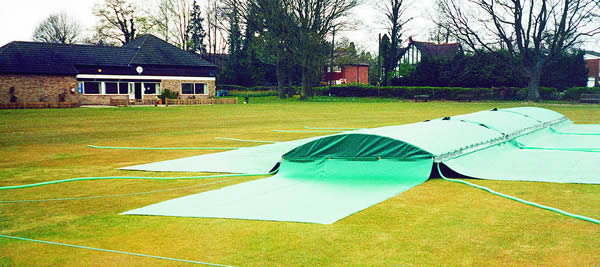 A cricket pitch covered by green protective covers on a grassy field, with a clubhouse and trees in the background under an overcast sky.
