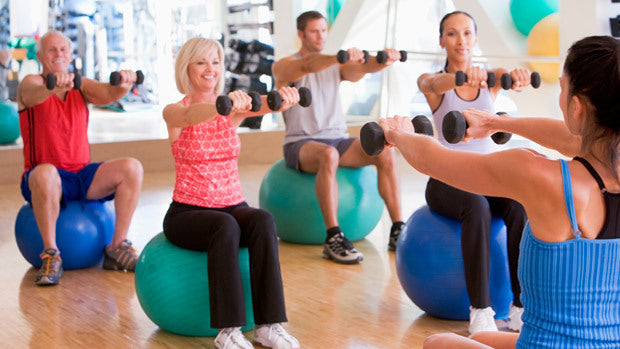 Five adults sit on exercise balls in a gym, holding dumbbells with arms extended forward, participating in a fitness class. They are dressed in athletic wear and smiling.
