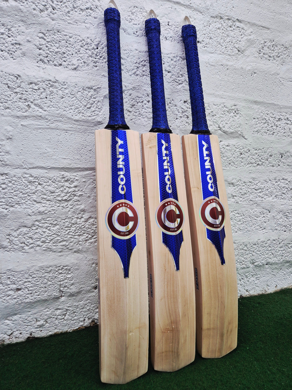 Three Hunts County Reflex Senior Cricket Bats, each with blue handles and COUNTY branding, stand upright against a white brick wall on a green surface—perfect for senior cricket players.