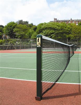 A close-up view of a tennis net on an outdoor tennis court with green and red surfaces, surrounded by trees and buildings in the background.