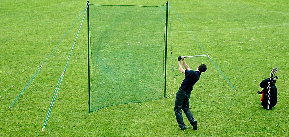 A person swings a golf club, hitting a ball into a practice net set up on a grassy field. A golf bag stands nearby on the right.