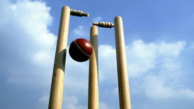 A cricket ball passing between three wooden stumps, dislodging the bails, set against a blue sky with scattered clouds.