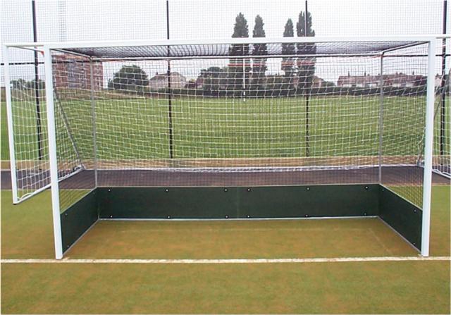 A field hockey goal with a net and a solid base stands on an outdoor artificial turf pitch, with green grass, trees, and buildings visible in the background.