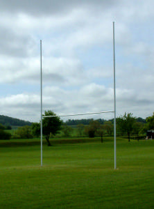 A rugby goalpost stands on a grassy field with trees and rolling hills in the background under a cloudy sky.