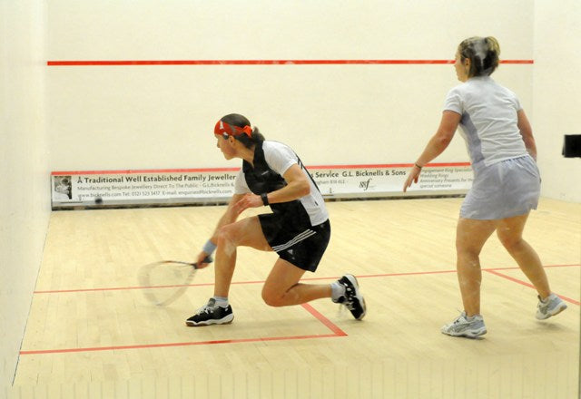 Two women play squash on an indoor court; one is lunging forward to hit the ball with her racket while the other stands behind her, watching and ready to react.