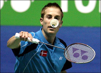 A badminton player in a blue shirt with a Denmark flag patch prepares to hit a shuttlecock, which is positioned in front of his face, during a match.