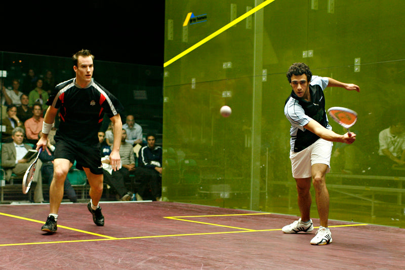 Two men playing squash on an indoor court, one preparing to hit the ball while the other watches, with spectators seated in the background.