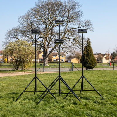 Three black tripod-mounted lights stand on a grassy field with houses and leafless trees in the background on a clear day.