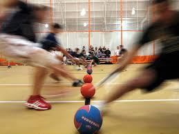 Blurry action shot of people running toward a line of dodgeballs in a gym, reaching to grab the balls as spectators watch from bleachers in the background.