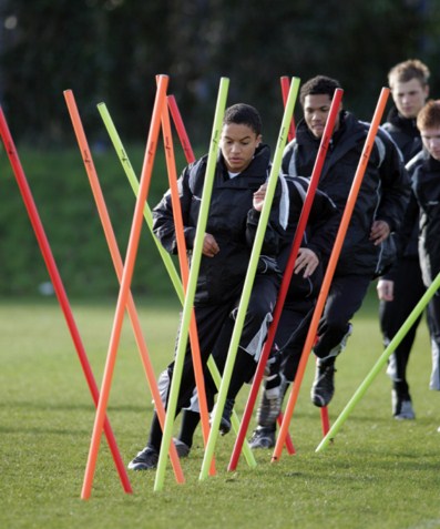 Four young athletes in black sportswear weave through brightly colored training poles on a grassy field, practicing agility drills. The focus is on the person leading the group.