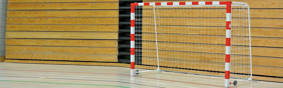 A handball goal with a red and white striped frame and net stands on a gymnasium floor with colored court lines and wooden panel walls in the background.