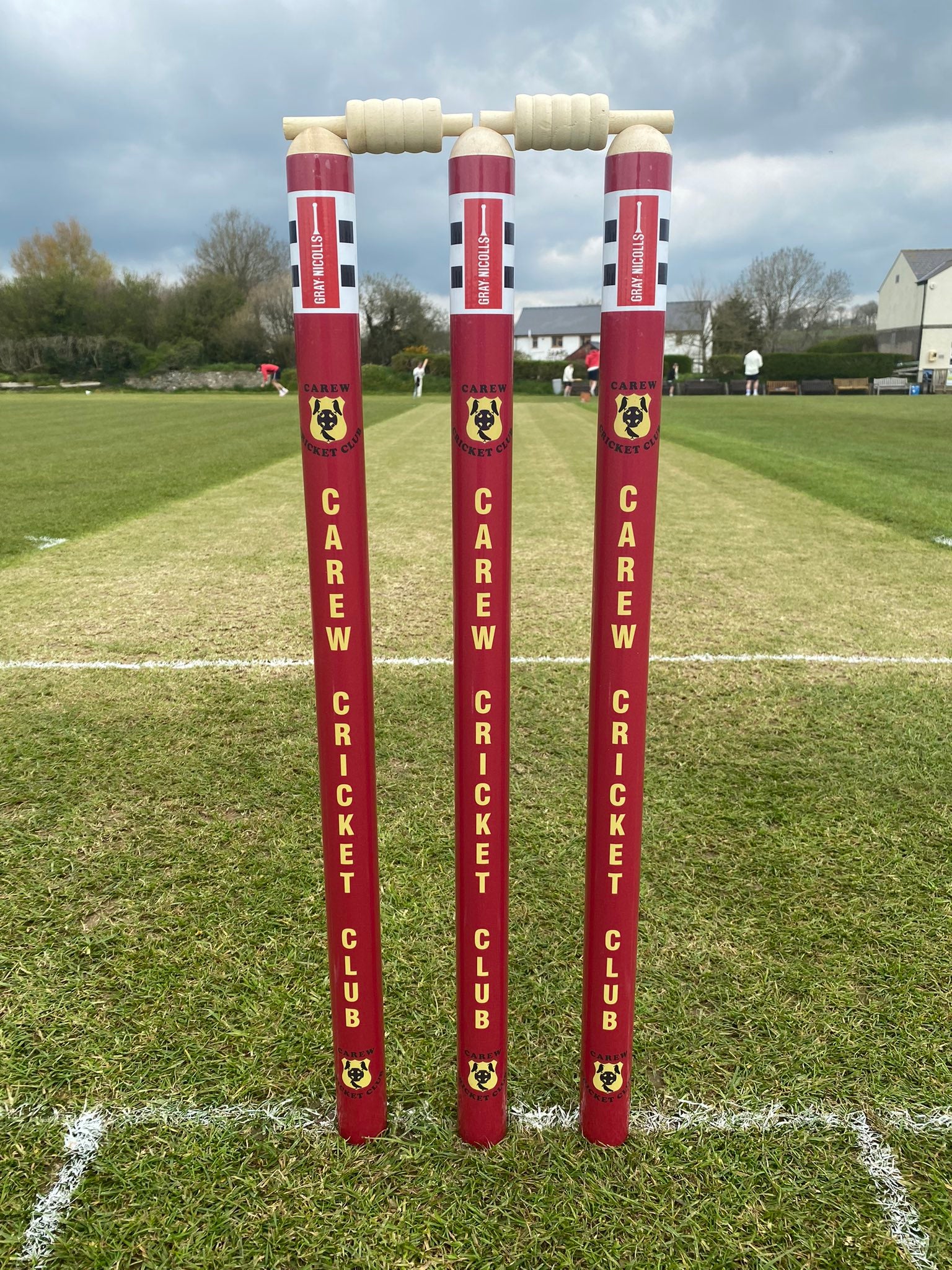 Three Gray Nicolls Custom Printed International Cricket Stumps, personalized with Carew Cricket Club and its logo, stand on a green field with bails on top. Behind them are trees, buildings by the field, and a cloudy sky.