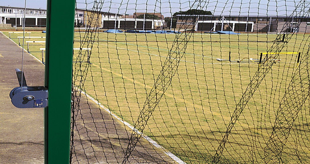 A sports field with hurdles and blue mats is seen through a net fence and a green metal post with a lock. School buildings are visible in the background under a clear sky.