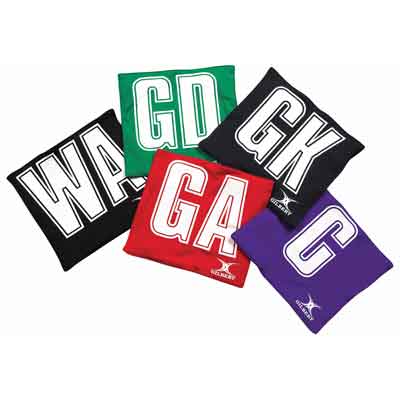 Five colorful netball bibs labeled WA, GD, GK, GA, and C are arranged overlapping each other. The bibs are green, black, red, purple, and black with bold white letters.