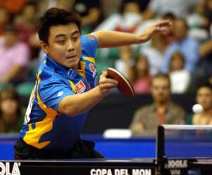 A table tennis player in a blue and yellow jersey lunges forward to hit the ball with his paddle during a match, with a blurred audience in the background.