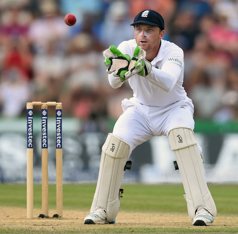 An England cricket wicketkeeper in white uniform and gloves crouches behind the stumps, ready to catch the ball during a match. The pitch and blurred spectators are visible in the background.