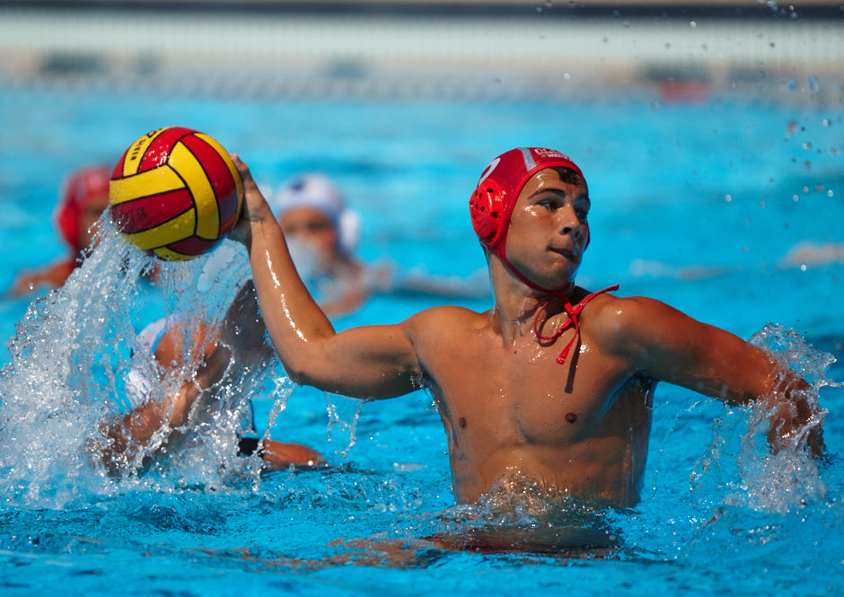 A male water polo player wearing a red cap prepares to throw the ball during a match in a pool, with other players visible in the background.