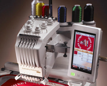 A computerized embroidery machine with colorful thread spools and a touchscreen displaying a red design that reads SWEET TREAT. The machine is stitching text on red fabric.
