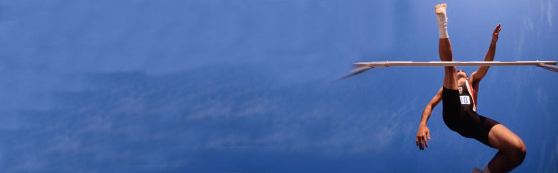 Athlete in midair clearing a high jump bar against a blue background, with arms and legs extended and one foot wrapped in athletic tape.