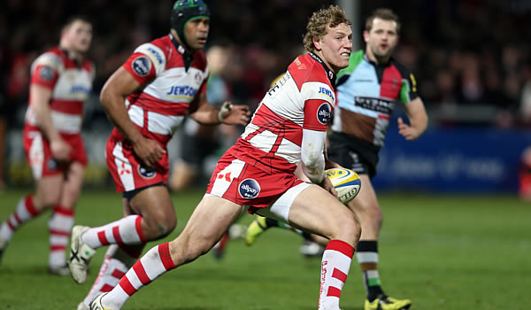 A rugby player in a red and white striped uniform prepares to pass the ball during a match, with teammates and opponents running alongside him on the field.