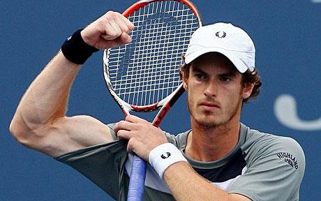 A male tennis player wearing a white cap and gray shirt holds a tennis racket and flexes his right arm in a victory gesture on an outdoor court.