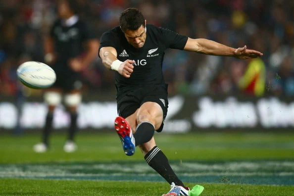 A rugby player in a black uniform kicks a ball during a match, with his right leg extended forward and a blurred crowd visible in the background.