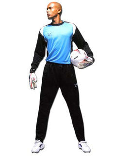 A male soccer goalkeeper in a blue and black uniform stands holding a soccer ball under one arm and wearing gloves, looking to the side against a white background.