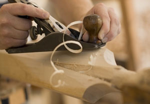 Close-up of hands using a hand plane to smooth a piece of wood, with wood shavings curling away from the tool.