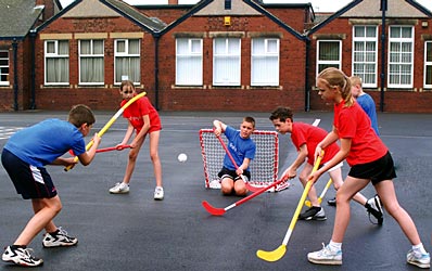 Six children play a game of street hockey on a playground; three in blue shirts and three in red. One child in blue acts as goalie, kneeling in front of a small red net as others compete for the ball.
