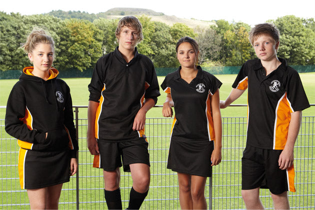 Four teenagers wearing black and orange sports uniforms stand in front of a wire fence on a grassy sports field, with trees and a hill in the background.
