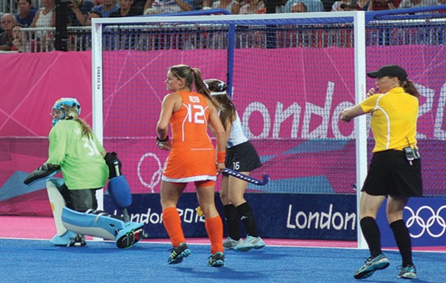 Field hockey players compete in front of a goal at the London Olympics; a player in orange, a goalkeeper in green, a player in dark blue, and a referee in yellow are visible on the blue field.