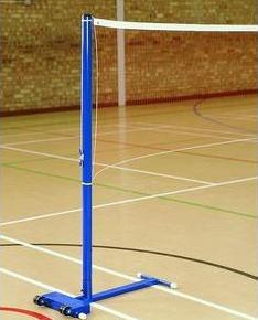 A blue badminton net post stands on a gymnasium floor with painted court lines and a brick wall in the background.