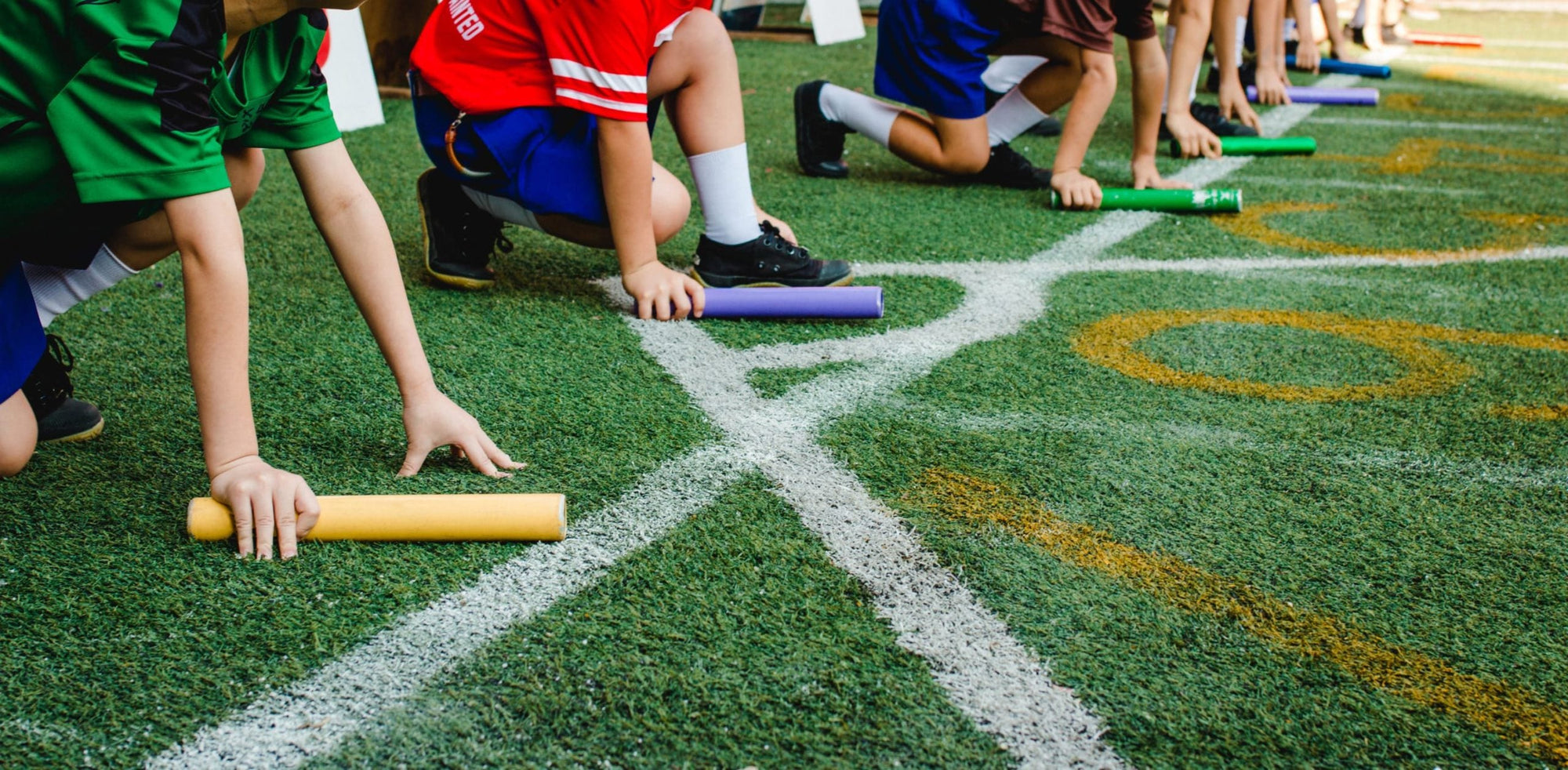 Children in sports uniforms crouch at a starting line on a field, each holding a different colored baton, preparing to start a relay race. Only their legs and arms are visible.