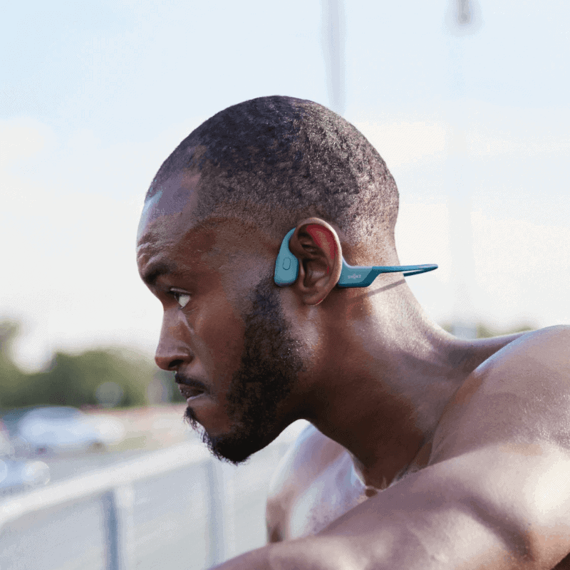 A man with a shaved head and beard wears teal bone conduction headphones outdoors, looking focused while exercising shirtless.