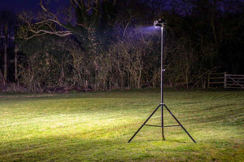 A tall tripod-mounted floodlight shines brightly on a grassy field at night, with leafless trees and a wooden gate visible in the background.