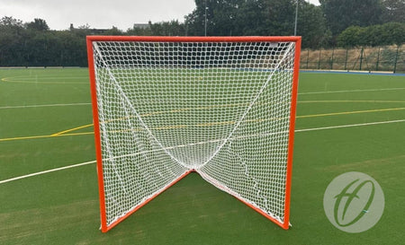 A Harrod Sport Freestanding Competition Lacrosse Goal, featuring a red frame and white netting, stands on green artificial turf marked with white and yellow lines, surrounded by trees in the background—ideal for regulation play.