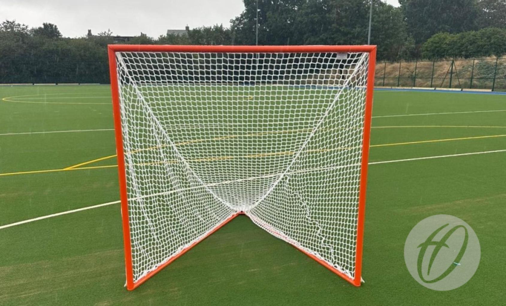 A Harrod Sport Freestanding Competition Lacrosse Goal, featuring a red frame and white netting, stands on green artificial turf marked with white and yellow lines, surrounded by trees in the background—ideal for regulation play.
