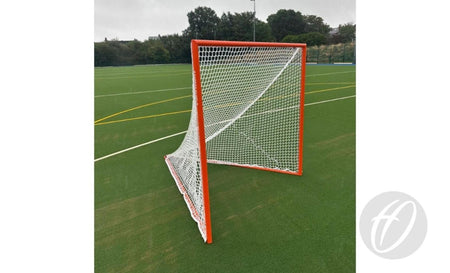 A Harrod Sport Freestanding Competition Lacrosse Goal, featuring a white net and orange frame, stands on a green artificial turf field marked with yellow and white lines, ready for play. In the background, trees and a fence are visible under a cloudy sky.