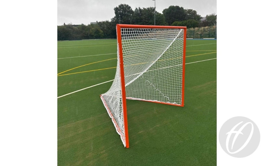 A Harrod Sport Freestanding Competition Lacrosse Goal, featuring a white net and orange frame, stands on a green artificial turf field marked with yellow and white lines, ready for play. In the background, trees and a fence are visible under a cloudy sky.