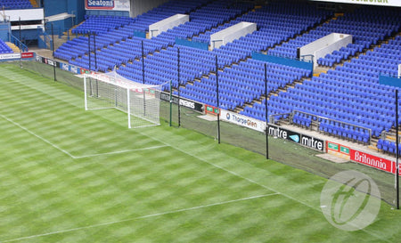 A Harrod Sport Crowd Protection Ball Stop System (100m x 5m) stands on a green field in an empty stadium, featuring sturdy aluminium poles, mesh netting behind the goal area, and rows of blue seats in the background.