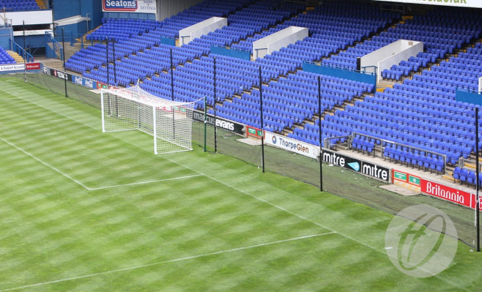 A Harrod Sport Crowd Protection Ball Stop System (100m x 5m) stands on a green field in an empty stadium, featuring sturdy aluminium poles, mesh netting behind the goal area, and rows of blue seats in the background.