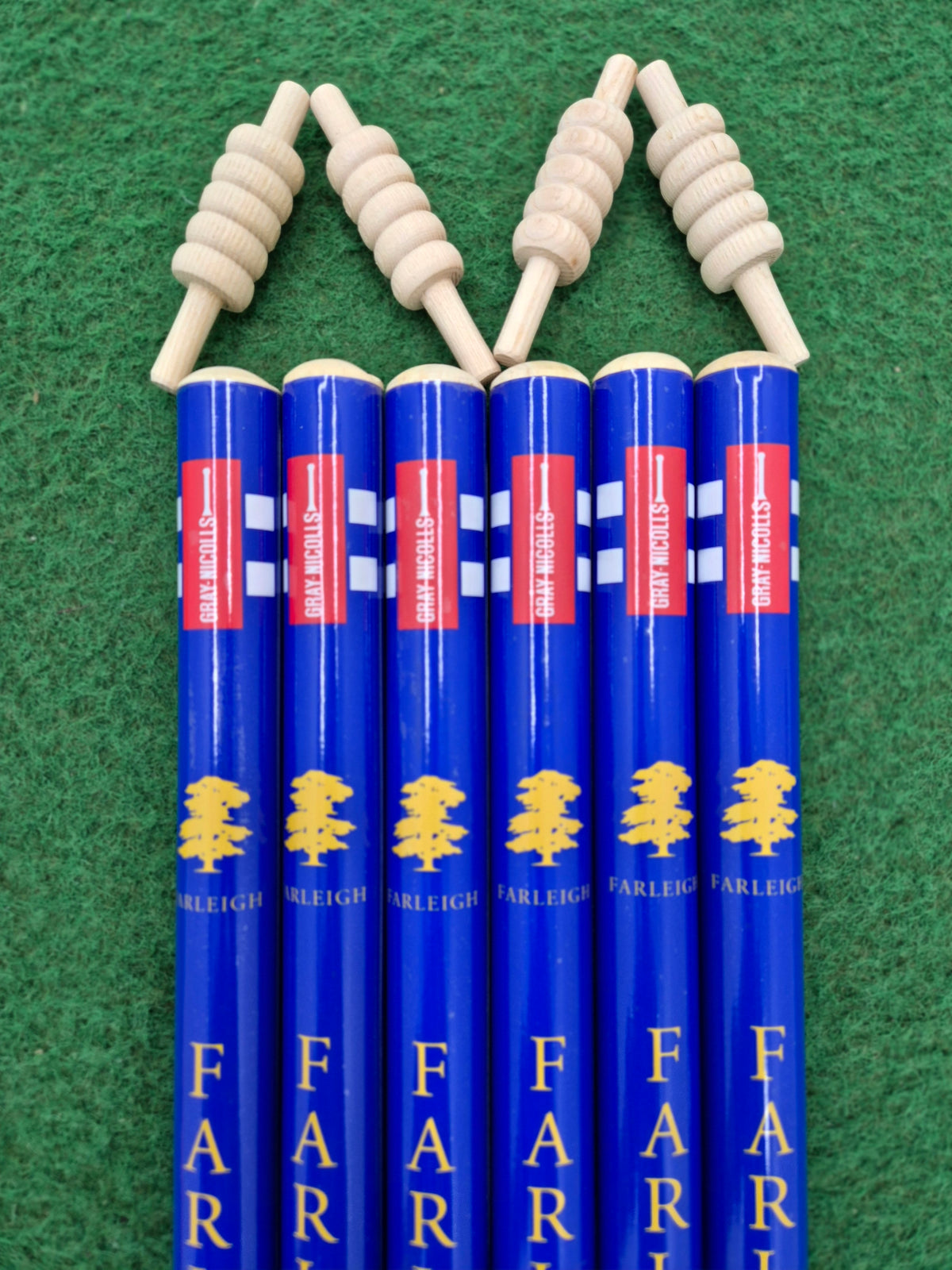 Six Gray Nicolls Custom Printed Club Cricket Stumps, each with blue finish, yellow tree logos, and "FARLEIGH" printed on them, stand vertically on green felt. Each stump is topped with a spiral-shaped wooden object, echoing cricket stumps.