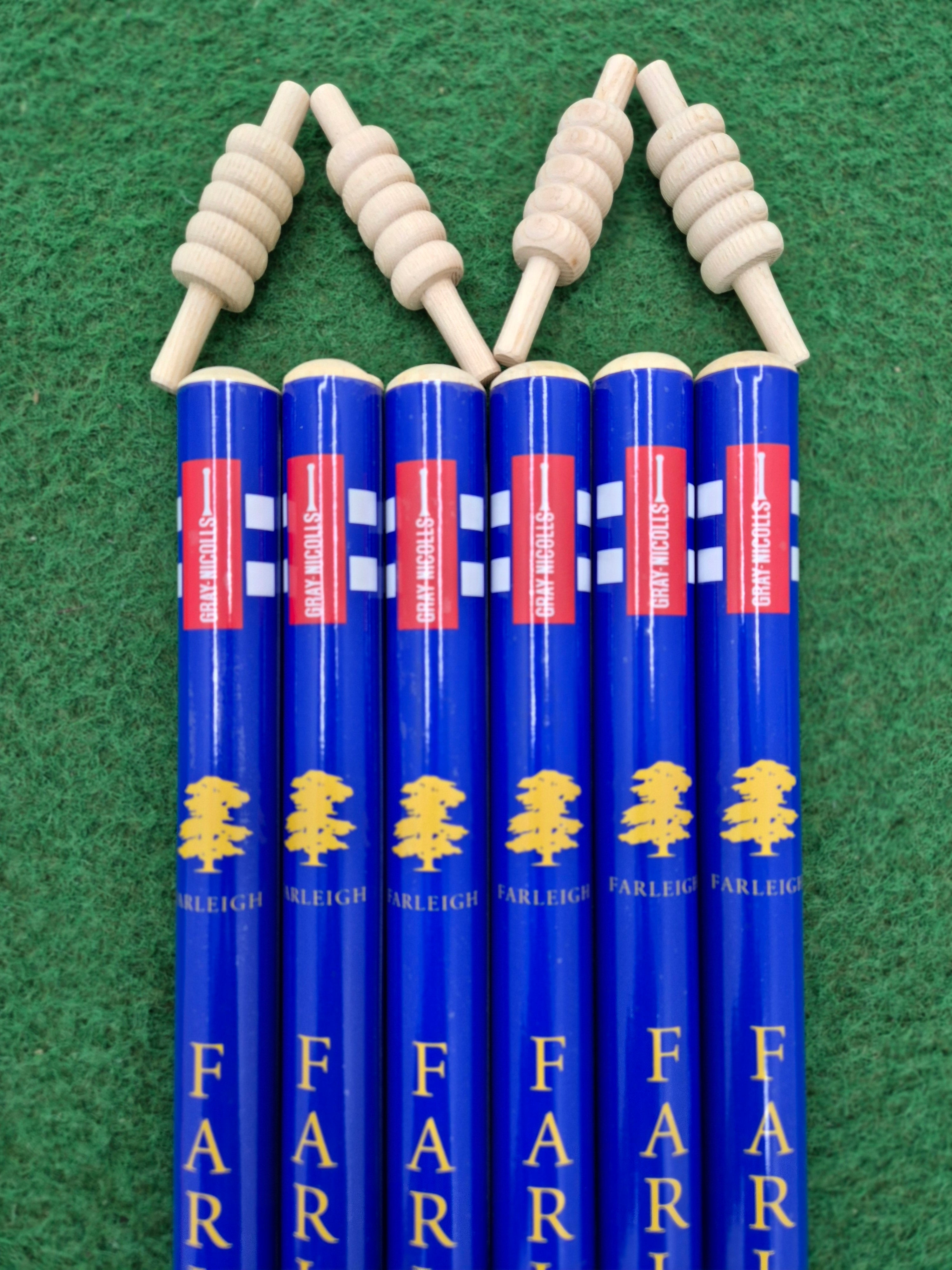Six Gray Nicolls Custom Printed Club Cricket Stumps, each with blue finish, yellow tree logos, and "FARLEIGH" printed on them, stand vertically on green felt. Each stump is topped with a spiral-shaped wooden object, echoing cricket stumps.