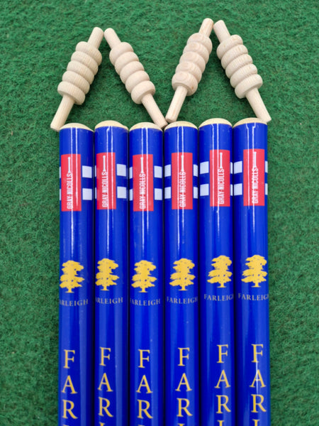 Six Gray Nicolls Custom Printed Club Cricket Stumps, each with blue finish, yellow tree logos, and "FARLEIGH" printed on them, stand vertically on green felt. Each stump is topped with a spiral-shaped wooden object, echoing cricket stumps.