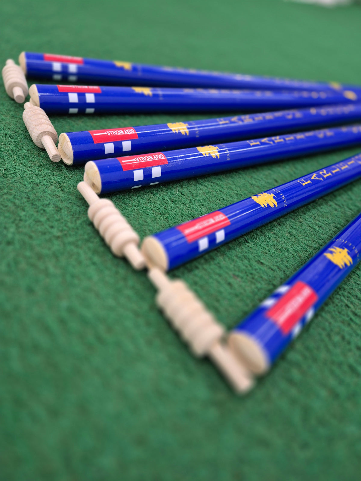 Six Custom Printed Club Cricket Stumps by Gray Nicolls are arranged diagonally on a green textured surface, with the ends facing the camera and displaying red, white, and yellow markings.