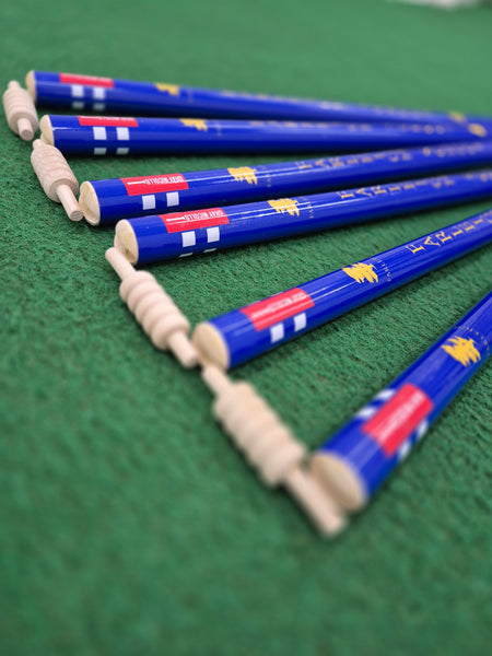 Six Custom Printed Club Cricket Stumps by Gray Nicolls are arranged diagonally on a green textured surface, with the ends facing the camera and displaying red, white, and yellow markings.