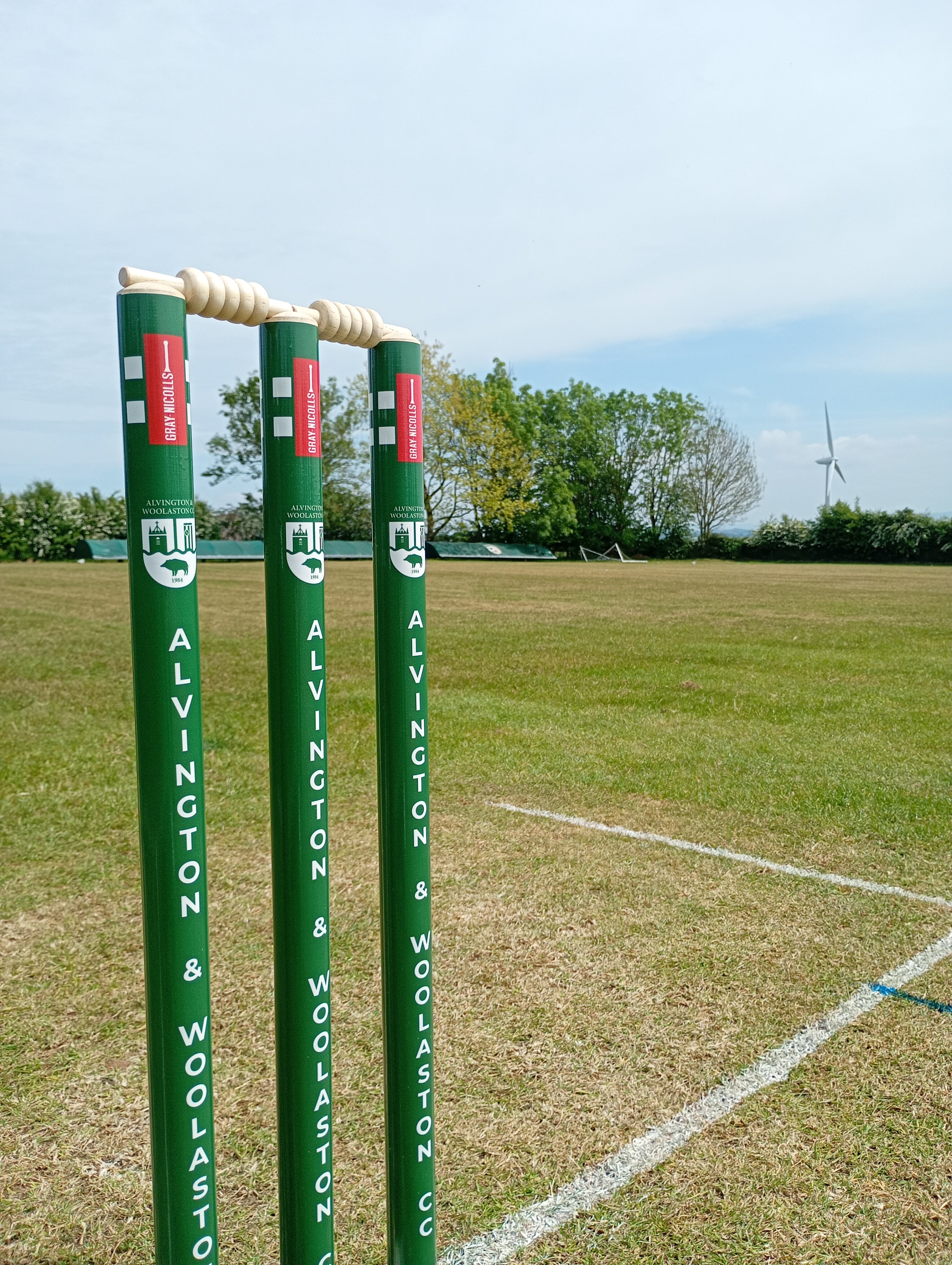 Three green Gray Nicolls Custom Printed Club Cricket Stumps with white bails stand on a grassy field marked by boundary lines, with trees and a wind turbine in the background under a partly cloudy sky.