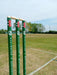 Three green Gray Nicolls Custom Printed Club Cricket Stumps with white bails stand on a grassy field marked by boundary lines, with trees and a wind turbine in the background under a partly cloudy sky.